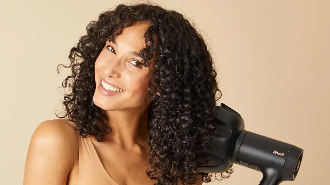 Smiling woman with curly hair holding a hairdryer.